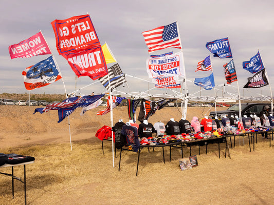 Busy campaign merchandise stand at a Trump rally, filled with a variety of political items including hats, flags, and shirts, with shoppers browsing and interacting with the merchandise.